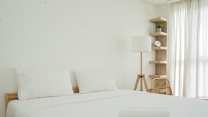 Ein ordentlich bezogenes Bett mit weißen Leinen und Kissen, eine Nachttischlampe, ein Holfnachttisch und ein kleines Regal mit Pflanze und Uhr.