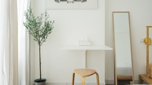 Ein schlichtes Badezimmer mit Blumentopf, einem Holzschemel, einem Spiegel und einem gerahmten Bild an der Wand.