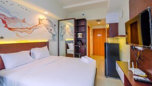 A hotel room with a large bed, a wall-mounted TV, a mini-fridge, a desk with a computer, and a decorative wall mural. - Restful and Tidy Studio at Evenciio Margonda Apartment (Depok)