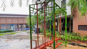 A playground with a red and green structure, surrounded by a wet pavement and greenery. - Simply Cozy 1BR at Asatti Apartment (Pagedangan)