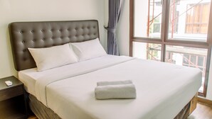 A neatly made bed with a tufted headboard, a bedside table with a phone and a book, and a window with curtains. - Simply Cozy 1BR at Asatti Apartment (Pagedangan)