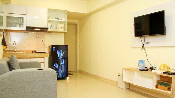 A compact living space with a kitchenette, a flat-screen TV mounted on the wall, a grey sofa, and a small wooden TV stand with electronic devices. - Cozy Living and Tidy 1BR Meikarta Apartment (Cikarang)