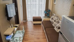 A compact living space with a sofa, a small table with a decorative glass top, a television mounted on the wall, a standing fan, and a refrigerator. - Nice and Spacious 2BR at Serpong Greenview Apartment (South Tangerang)
