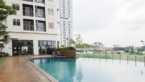 A swimming pool with a tiled deck, a building with multiple floors and balconies, and a view of a cityscape in the background. - Nice and Spacious 2BR at Serpong Greenview Apartment (South Tangerang)