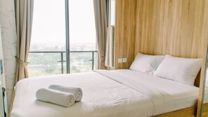 A neatly made bed with white linens and two rolled towels, a wooden headboard, and a view of a cityscape through large windows. - Cozy and Simply Studio at Sky House BSD Apartment (Pagedangan)