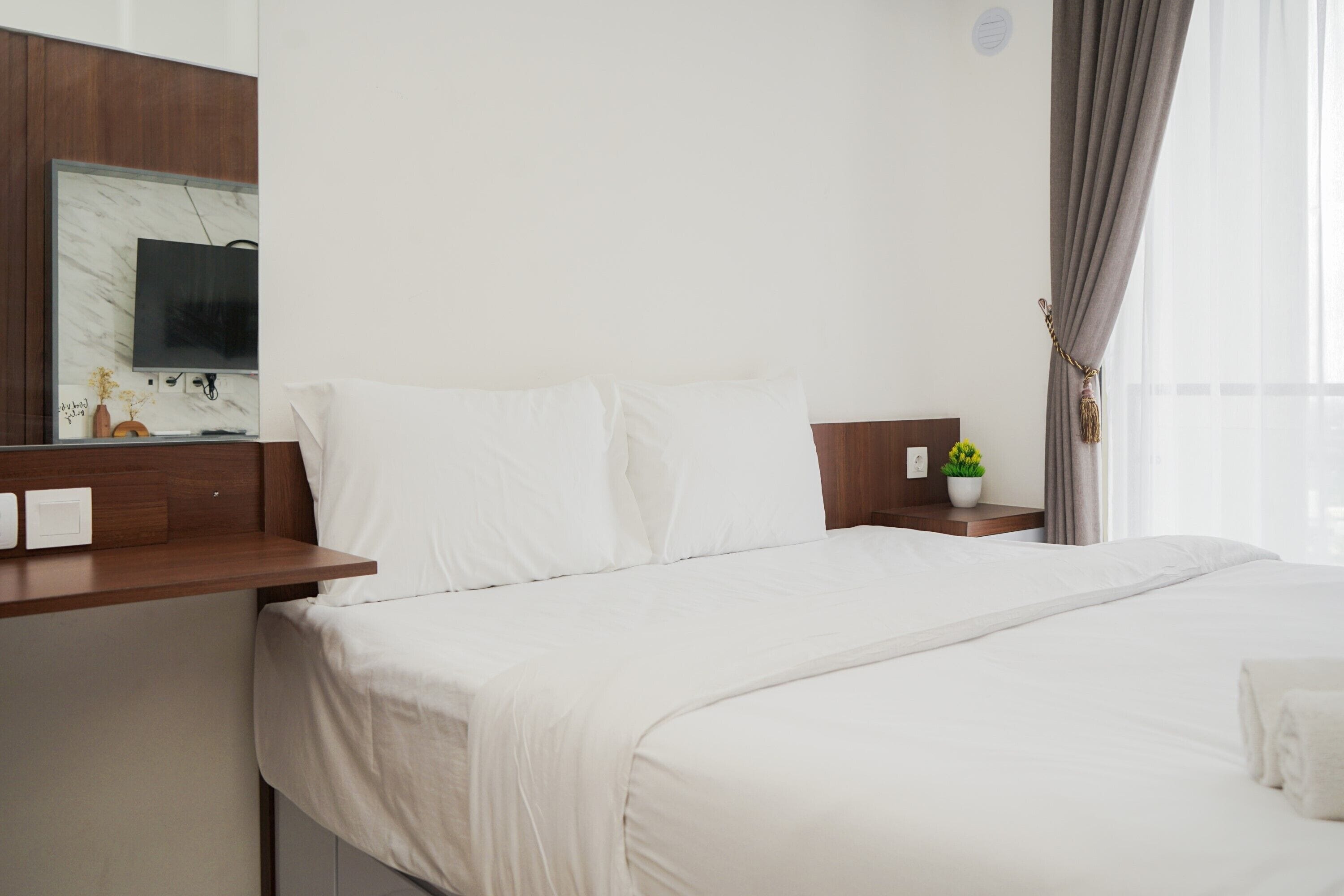 A hotel room with a large bed, a wooden nightstand, a flat-screen TV mounted on the wall, and a window with curtains.