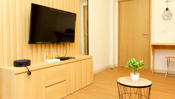 A modern room with a flat-screen TV mounted on a wooden wall, a small table with a potted plant, and a wooden door in the background. - Golden Osaka 2BR Apartment at Meikarta Apartment (Cikarang)