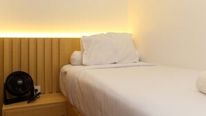 A neatly made bed with white bedding and pillows, a wooden headboard, and a small bedside table with a fan and a power outlet. - Golden Osaka 2BR Apartment at Meikarta Apartment (Cikarang)