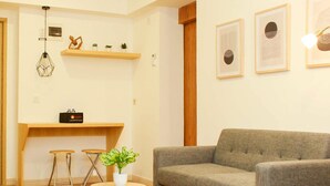 A living room with a grey sofa, a wooden dining table with two stools, a small round table with a plant, and a wall-mounted shelf with decorative items. - Golden Osaka 2BR Apartment at Meikarta Apartment (Cikarang)