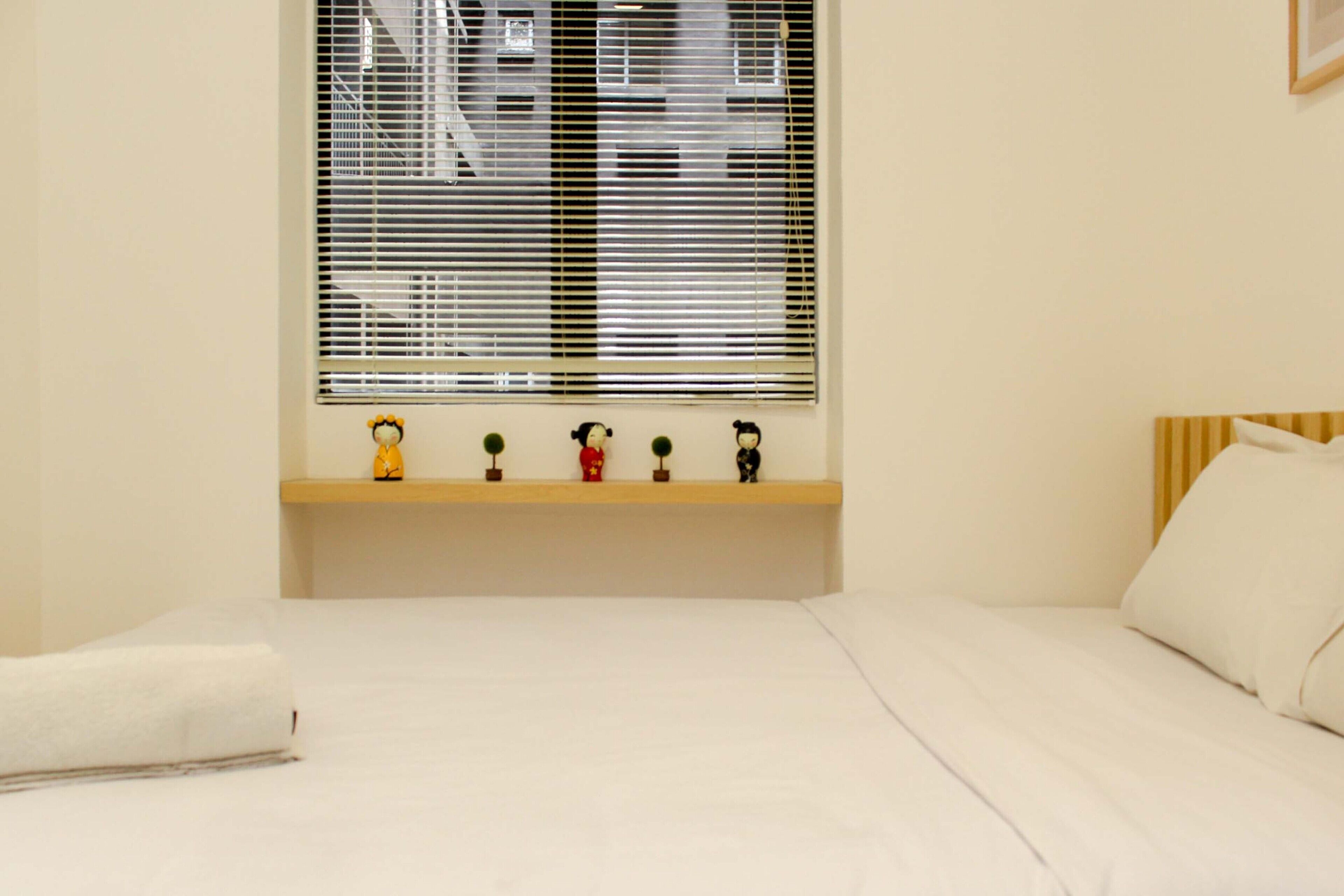 Een slaapkamer met een bed, een raam met jaloezieën, een plank met decoratieve objecten en een kussen.