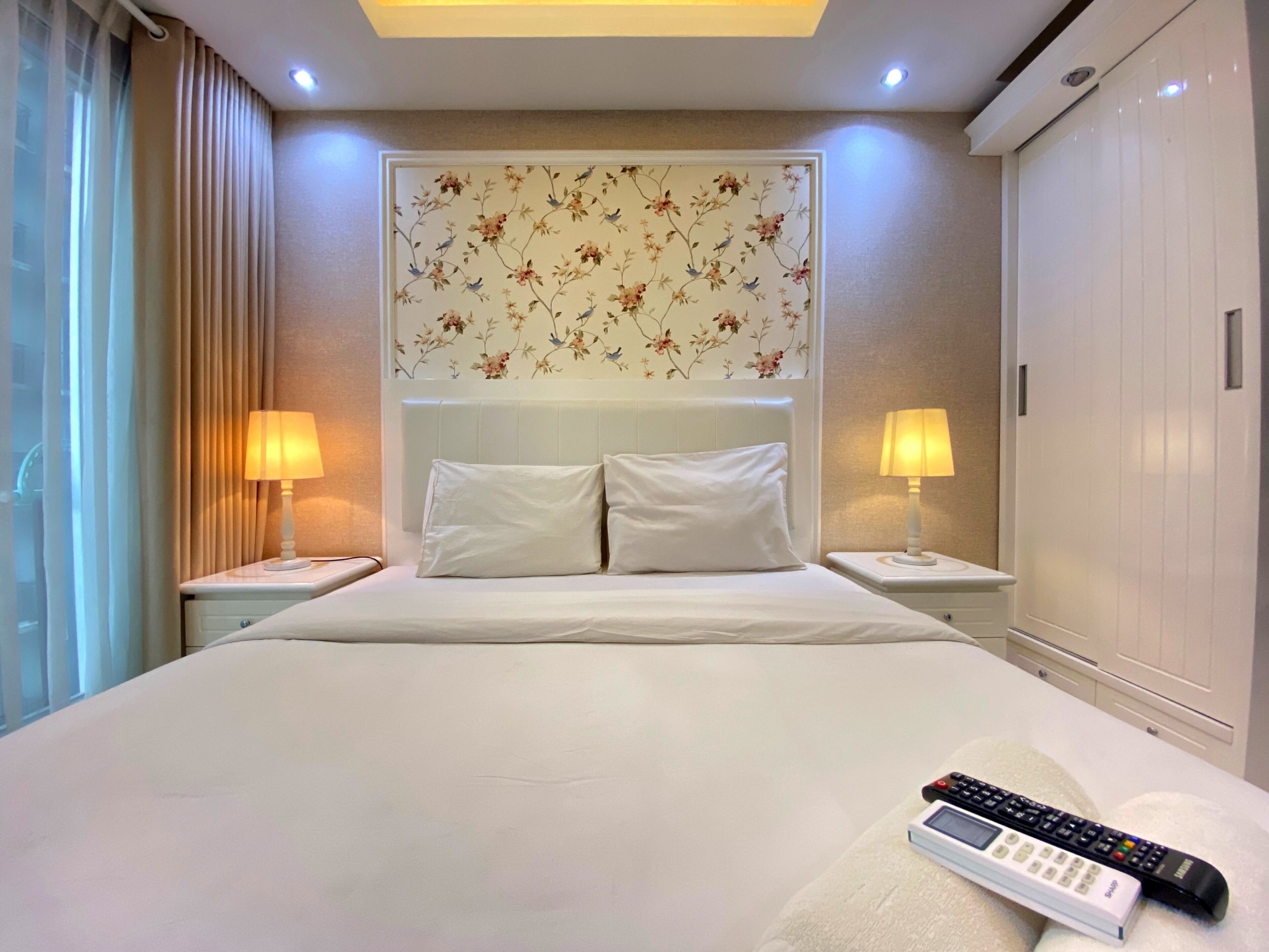 Une chambre d’hôtel avec un grand lit, deux lampes de chevet, une tête de lit recouverte d’un papier peint à motifs floraux et une télécommande posée sur le lit.
