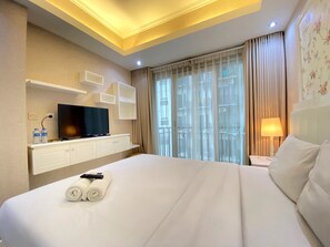A hotel room with a large bed, a flat-screen TV on a white cabinet, a window with curtains, and a bedside lamp. - Well Furnished Studio Room at Grand Asia Afrika Apartment (Bandung)