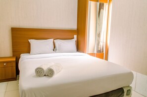 Una cama bien hecha con sábanas blancas y dos toallas enrolladas, un cabecero de madera y una mesita de noche con cajón.