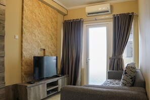 A living room with a flat-screen TV on a wooden stand, a grey sofa with patterned cushions, and a large window with curtains. - Comfort Living 3BR at Grand Palace Kemayoran Apartment (Jakarta)