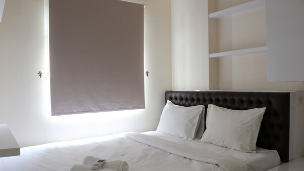 A neatly made bed with white linens and two pillows, a brown upholstered headboard, and a window with a brown blind.