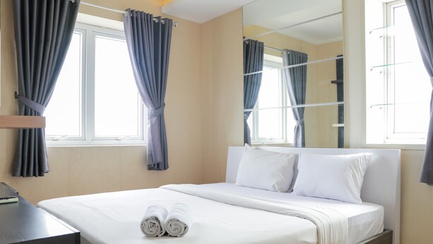 A neatly made bed with white linens, a bedside table with a remote control, and a large mirror on the wall. - Lavish 3BR Residence at Grand Palace Kemayoran (Jakarta)