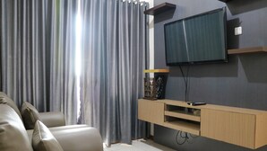 A modern living room with a flat-screen TV mounted on the wall, a wooden entertainment unit, and a grey sofa. - Lavish 3BR Residence at Grand Palace Kemayoran (Jakarta)