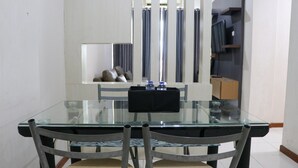 A dining area with a glass table, four chairs, a black tray with glasses, and a view of a living area with a TV and shelves. - Lavish 3BR Residence at Grand Palace Kemayoran (Jakarta)