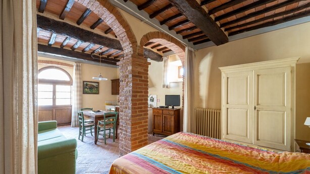 Une chambre avec un lit, une commode et une fenêtre. On aperçoit un coin repas avec une table et des chaises à travers une arche.