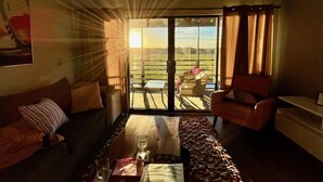 Een woonkamer met een bank, fauteuil en salontafel. Er is een groot raam met uitzicht op een veld en een gebouw.