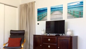 A hotel room with a bed, a wooden chair with a red cushion, a TV on a wooden cabinet, and three paintings of a pier on the wall.