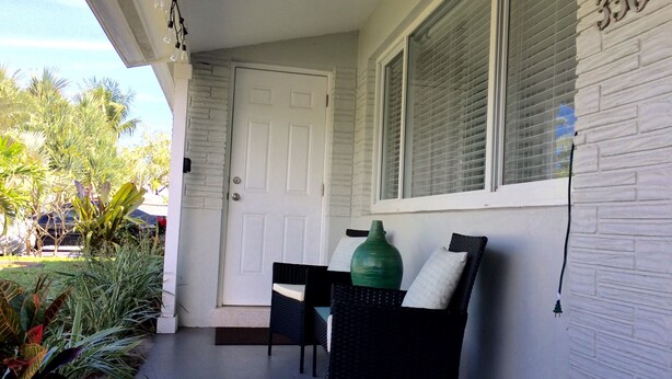 A clean, white-walled porch with a door, a window with blinds, and a green vase on a chair.