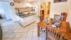 A modern kitchen with a dining area, featuring a large island, white cabinetry, and a dining table set for four. - Zen City Balcony ~ Outdoor Dining ~ Mindal Markets (Larrakeyah)