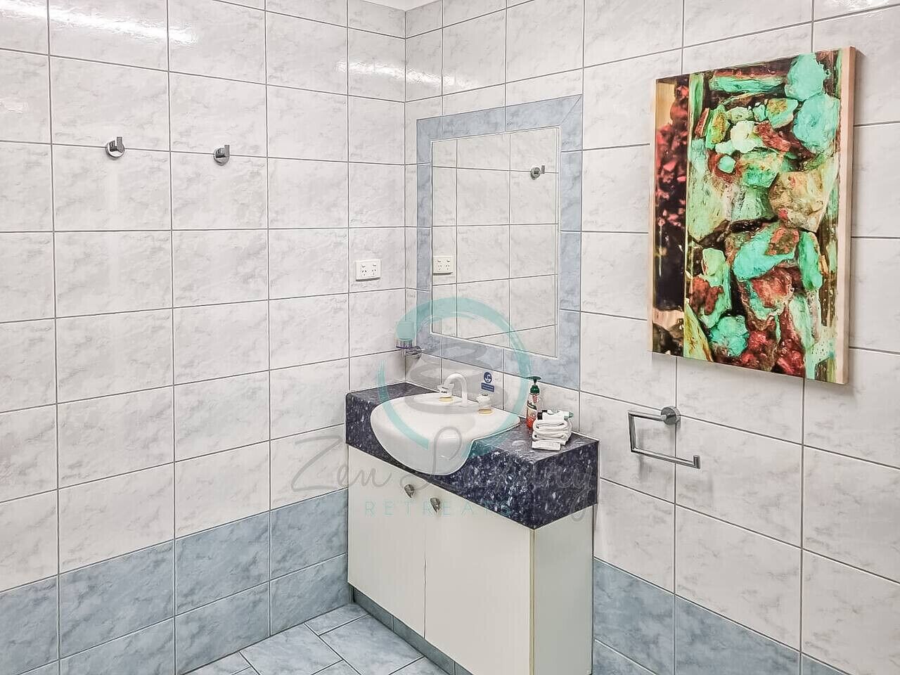 A bathroom with white marble tiles, a sink with a dark countertop, and a painting on the wall.