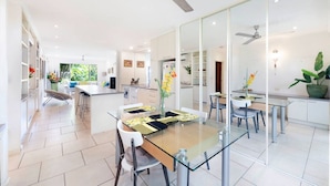 A modern kitchen with a central island, white cabinetry, and a dining area with glass tables and chairs. - Zen Treetops: Private Pool ~ Outdoor Dining (Larrakeyah)