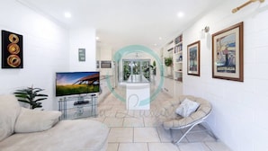 A modern living room with a TV, sofa, and armchair, a kitchen area with a dining table, and a view of the outdoors. - Zen Treetops: Private Pool ~ Outdoor Dining (Larrakeyah)