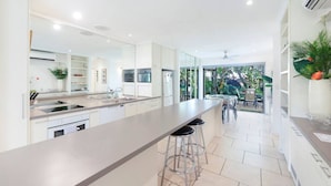 A modern kitchen with a central island, bar stools, and a dining area with a view of the garden. - Zen Treetops: Private Pool ~ Outdoor Dining (Larrakeyah)