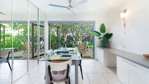 A modern kitchen with a glass dining table, a ceiling fan, and a view of the garden through large glass doors. - Zen Treetops: Private Pool ~ Outdoor Dining (Larrakeyah)