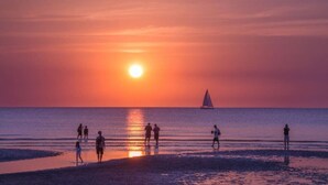 Um pôr do sol sobre uma praia com pessoas e um veleiro.