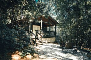 A cabin with a porch, wooden deck, and outdoor seating area surrounded by dense forest.