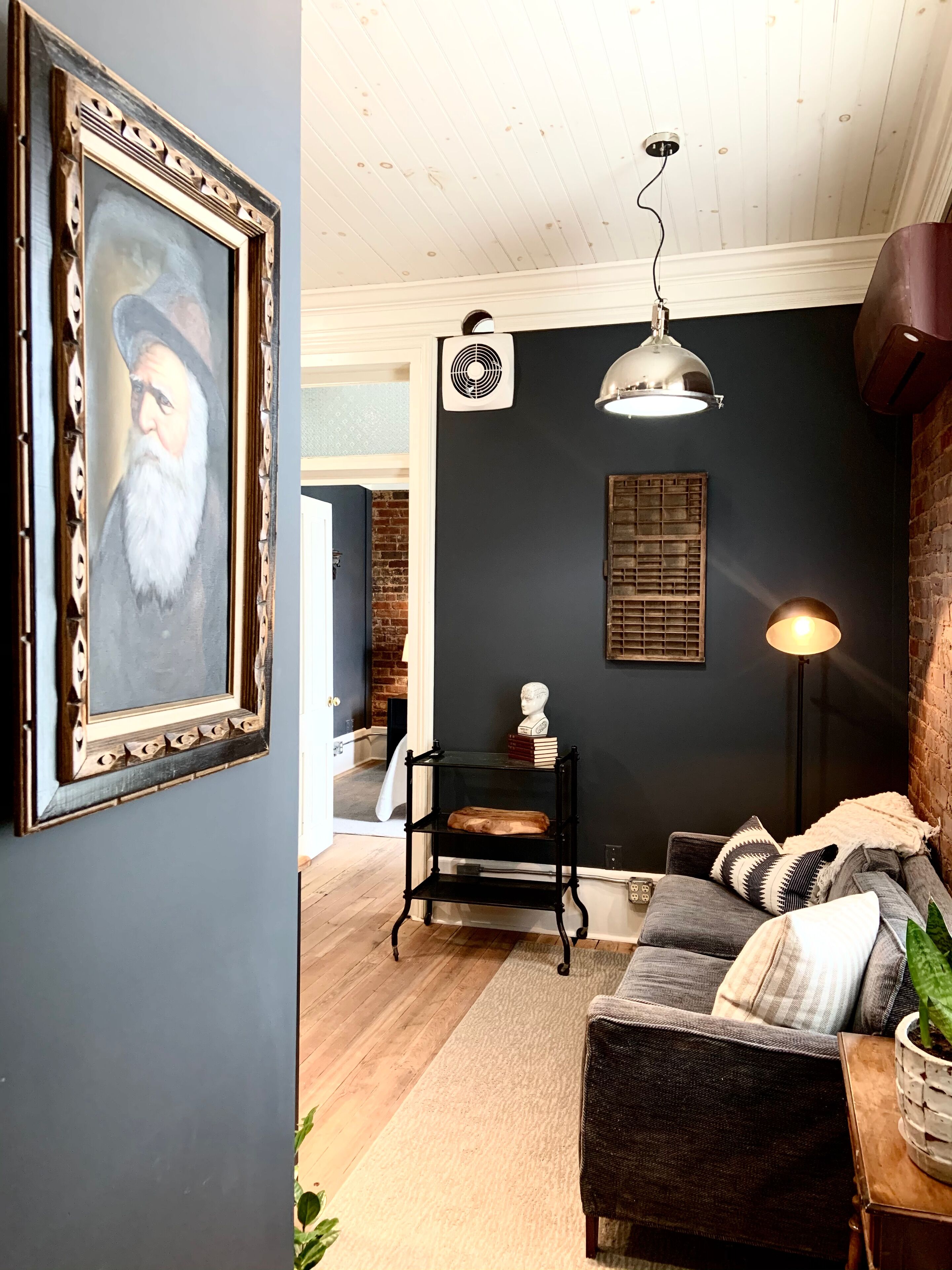 A cozy living room with a dark wall, a wooden floor, a black metal shelf with books, a white bust, and a lamp.