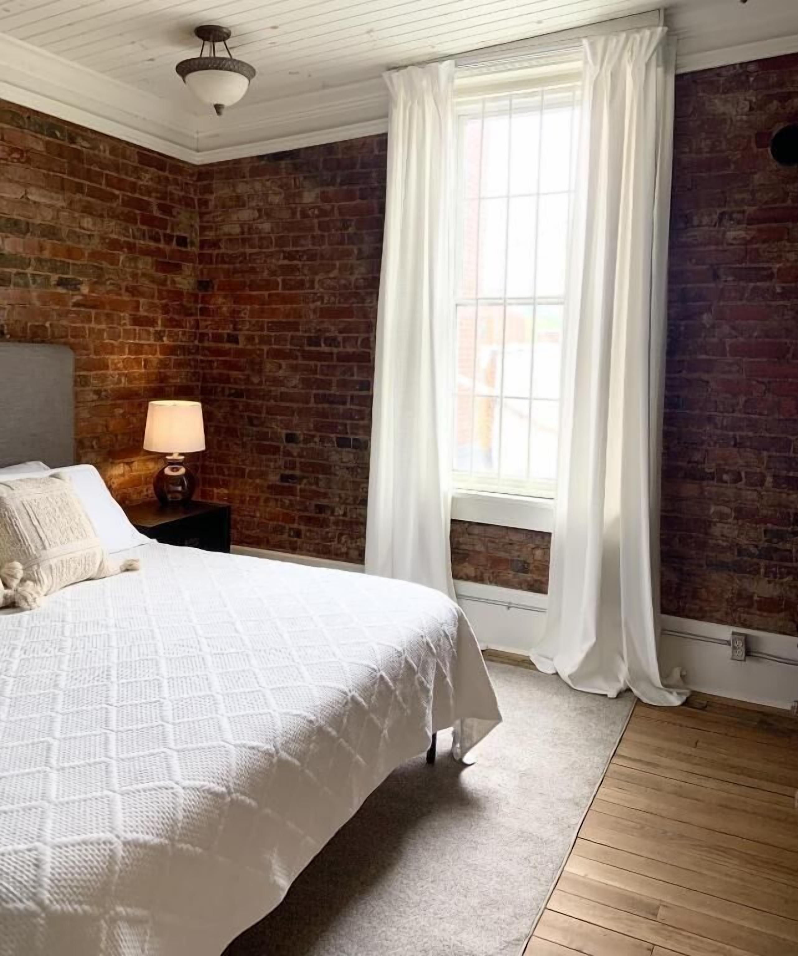 A bedroom with a brick wall, a wooden floor, a bed with a white bedspread, a nightstand with a lamp, and a window with white curtains.