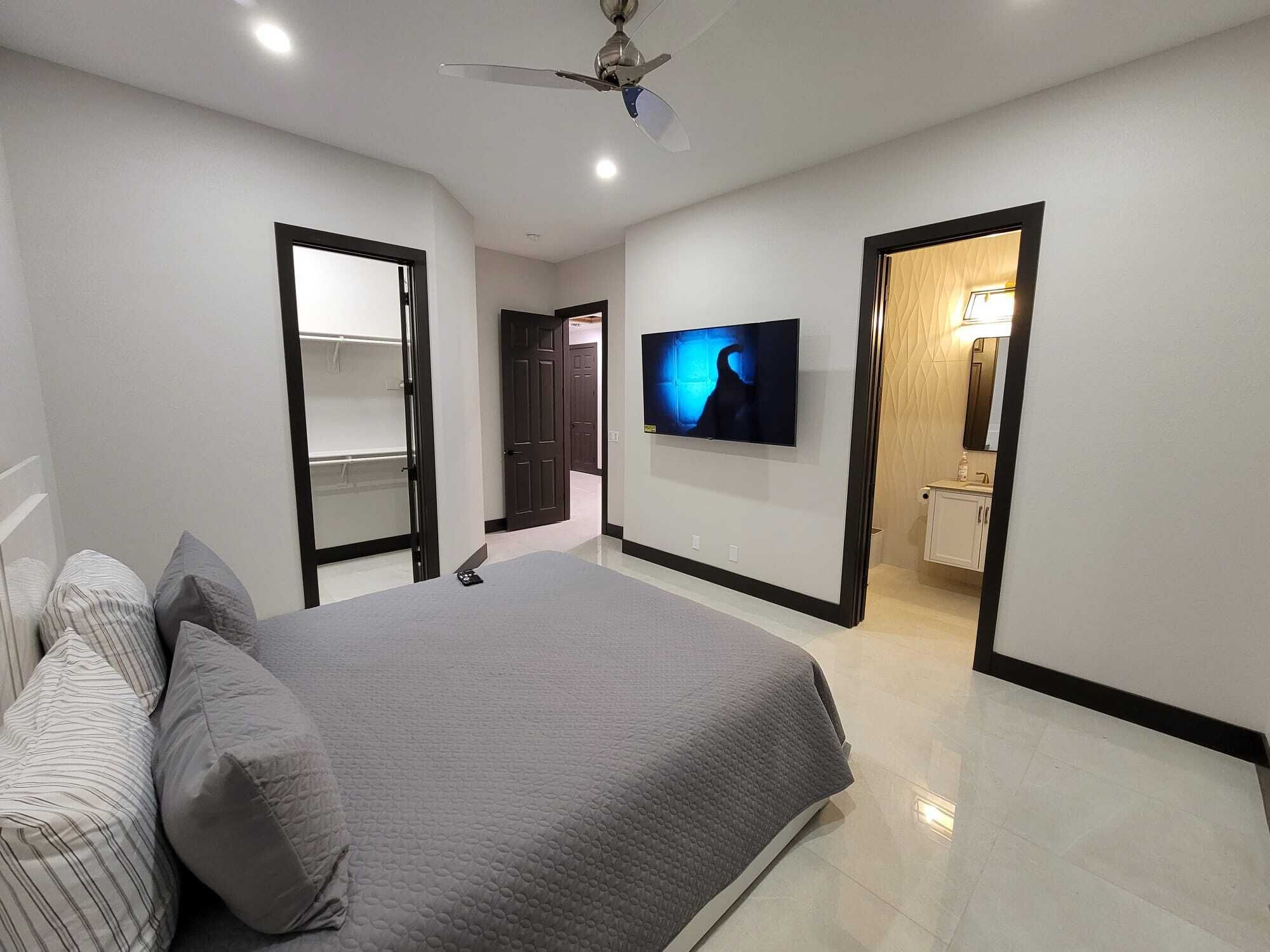 Un dormitorio con cama, televisión, ventilador de techo y baño visible a través de una puerta abierta.