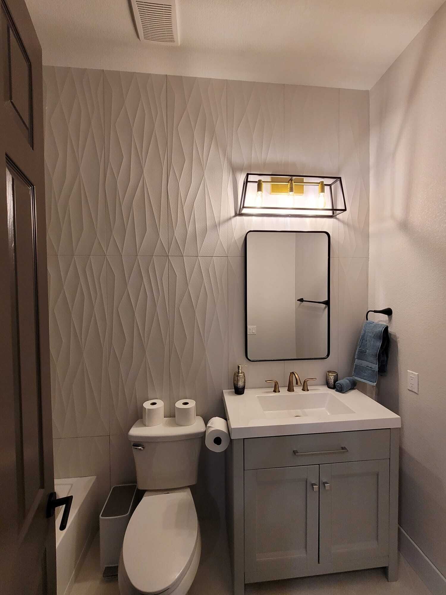 A modern bathroom with a white textured wall, a rectangular mirror, a white sink with a gold faucet, and a toilet with two rolls of toilet paper.