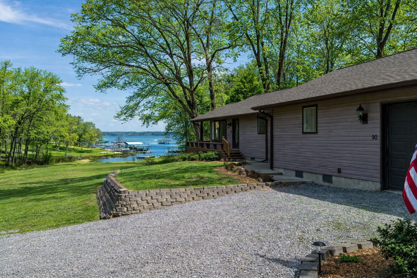 A single-story house with a gravel driveway, a wooden deck, and a view of a lake with boats.