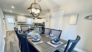A dining area with a wooden table set for six, surrounded by chairs, in a modern kitchen with white cabinets and a chandelier. - Lakefront Retreat with Dock and Fire Pit (Gilbertsville)