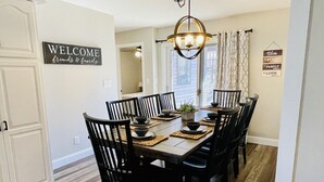Een eetkamer met een houten tafel voor zes personen, omringd door zwarte stoelen. Een bord met de tekst 'WELCOME friends & family' hangt aan de muur, en een bord met de tekst 'Blessed food family love' hangt boven de tafel.
