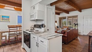 A compact kitchen with white cabinets, a stove, and a microwave. There is a dining table with chairs and a brown leather couch in the living area. - Fort Marcy 33 | Walk to Plaza | Kiva Fireplace | Full Kitchen | Hot Tub | Pool | Single Level (Santa Fe)