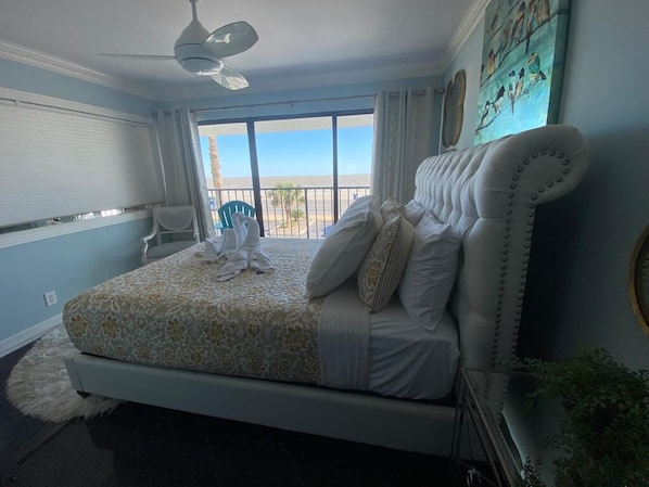 A bedroom with a large bed, a ceiling fan, and a view of the beach through the window.