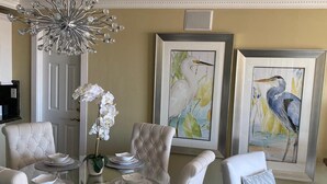 Un coin repas avec une table en verre entourée de chaises blanches à revêtement, des tableaux représentant des oiseaux encadrés au mur et un lustre au-dessus.