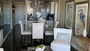 Un salon moderne avec une table basse en verre, des canapés blancs, un lustre et un coin repas avec une table et des chaises blanches.