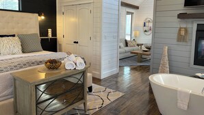 Una habitación de hotel moderna con cama, tocador con espejo, bañera y una zona de estar con sofá y mesa de centro.