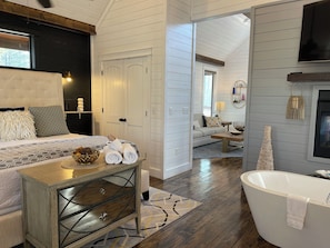 A modern hotel room with a bed, a vanity with a mirror, a bathtub, and a living area with a sofa and coffee table.