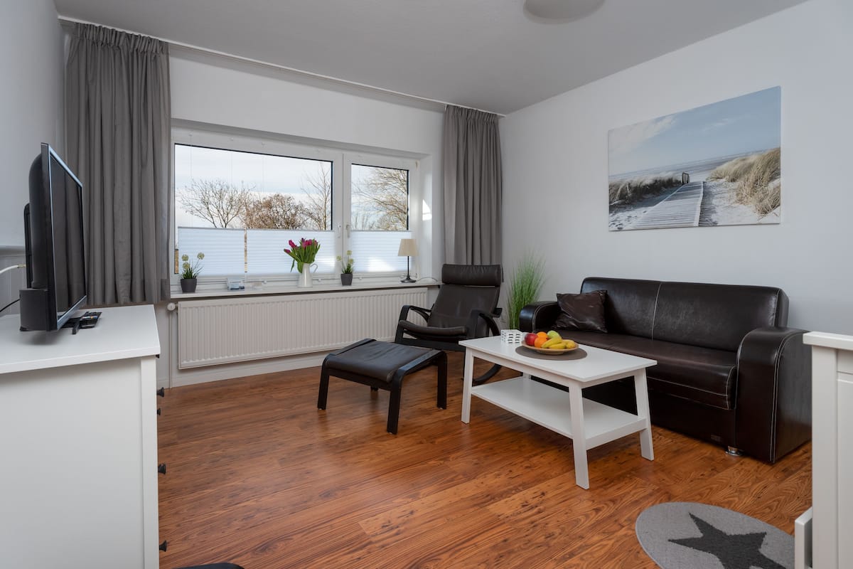 Un salon moderne avec un plancher en bois, une table basse blanche, un canapé en cuir marron et une grande fenêtre avec des rideaux.