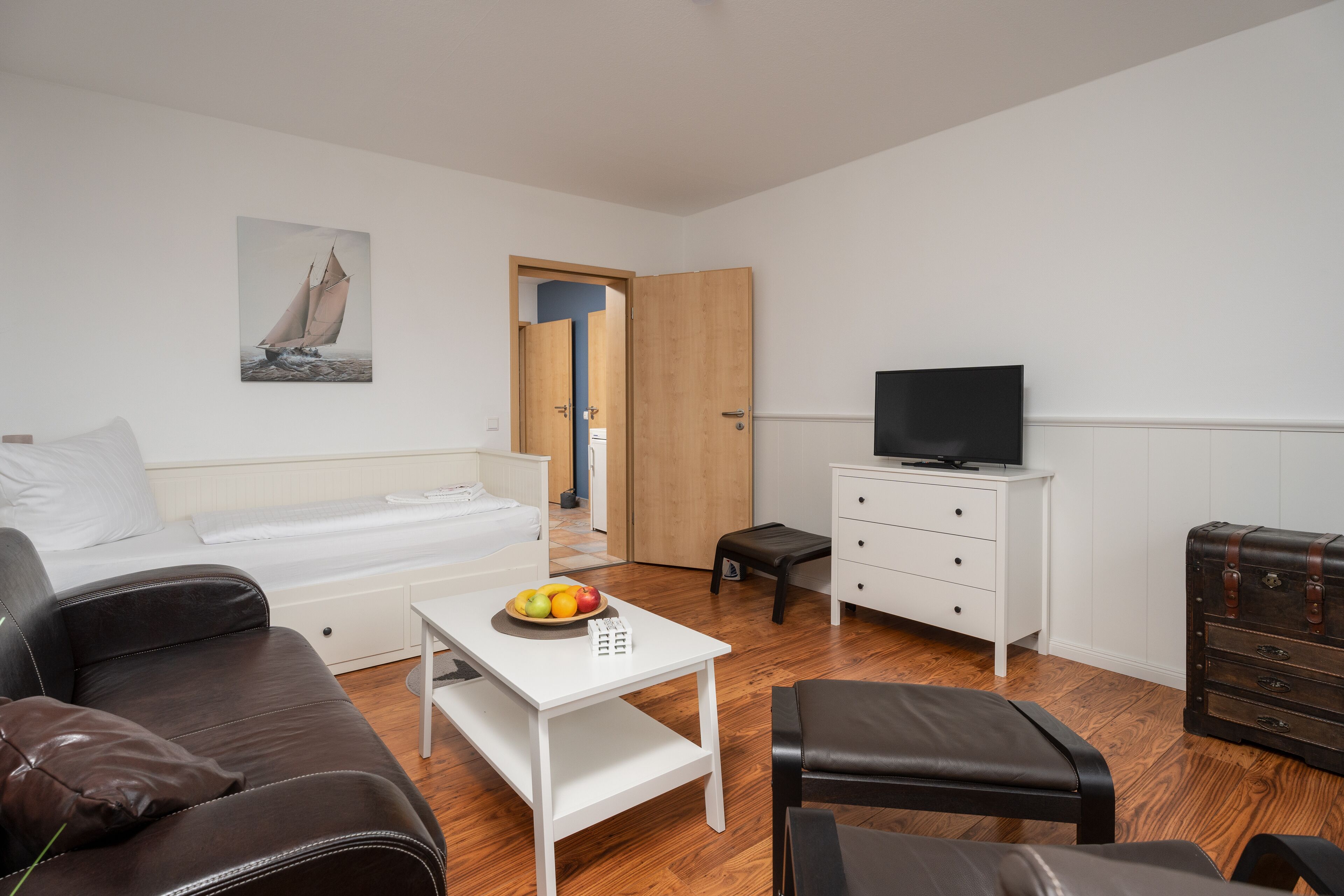 A modern hotel room with a bed, a white desk, a television, a leather sofa, and a suitcase.