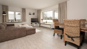 A modern living room with a sofa, dining table, wicker chairs, a TV, and large windows. - Ferienwohnungen Haus Schau ins Land (Emmelsbuell-Horsbuell)